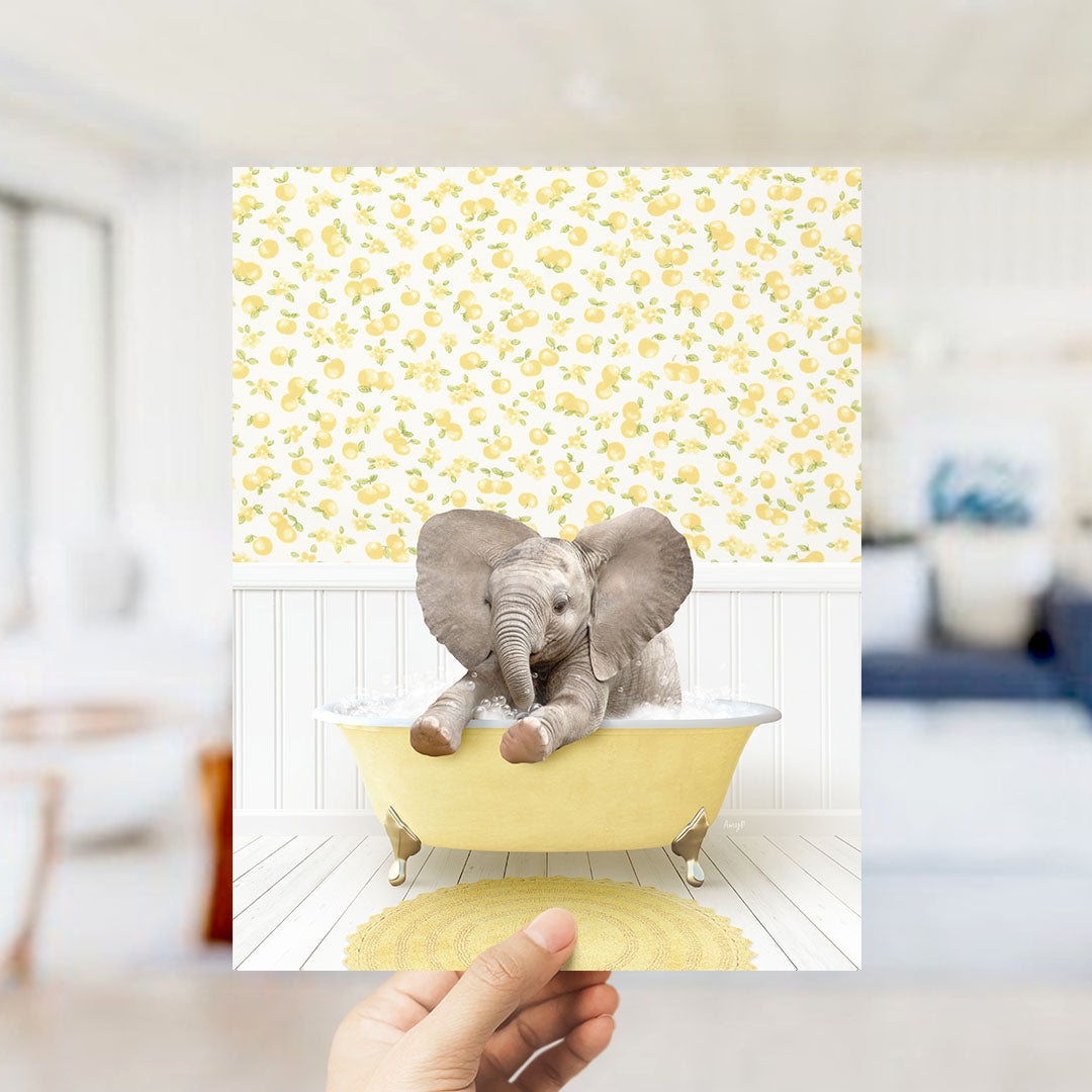 A hand holds up a framed image of a baby elephant sitting in a yellow bathtub, surrounded by a yellow and white floral pattern.