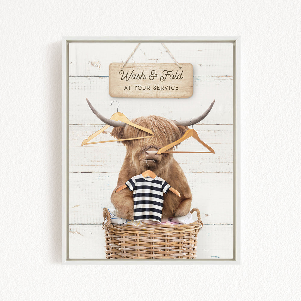 A framed image depicting a large, furry animal, possibly a yak or a yak dog, sitting in a wicker basket and holding a clothesline with clothes hanging on it. The animal appears to be in a relaxed and comfortable position, with a sign above it that reads "Wash & Fold at your Service".