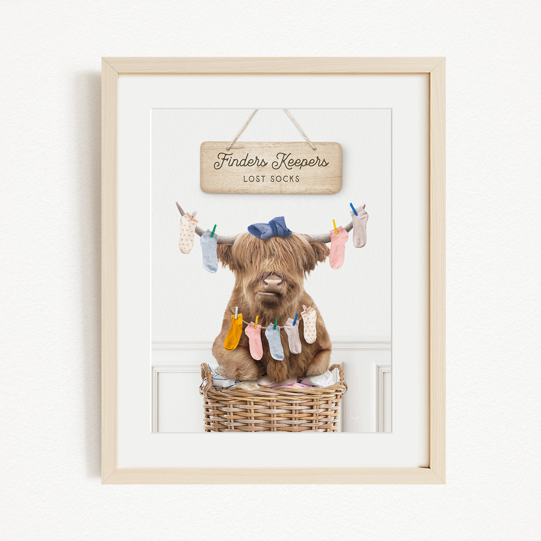 A framed image depicting a brown cow sitting in a wicker basket, with a sign above it that reads "Finders Keepers Lost Socks".