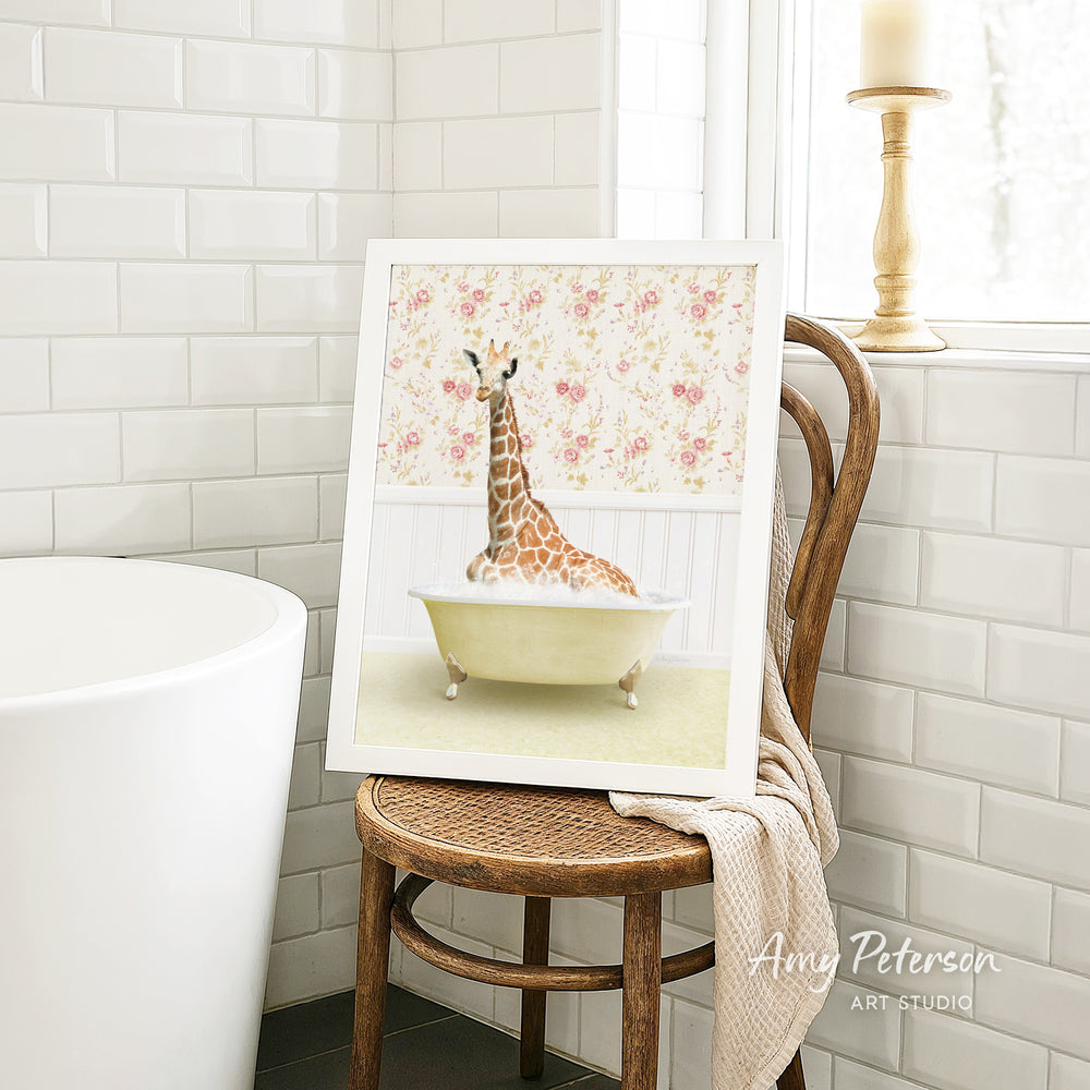 A framed image of a giraffe sitting in a bathtub is displayed on a wooden chair in a bathroom setting.