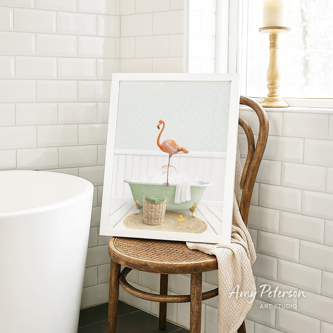 A framed image of a pink flamingo standing in a green bathtub, with a towel and basket nearby, is displayed on a wooden chair in a bathroom setting.