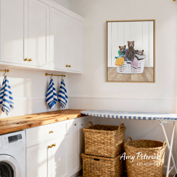 A framed image of three bears sitting in a laundry basket is hanging on a white wall in a bright, airy laundry room.