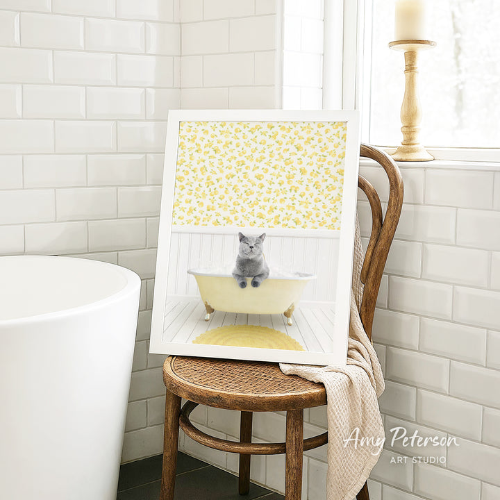 A framed image of a gray cat sitting in a yellow bathtub, with a yellow floral pattern on the tub and a wooden chair in the background.
