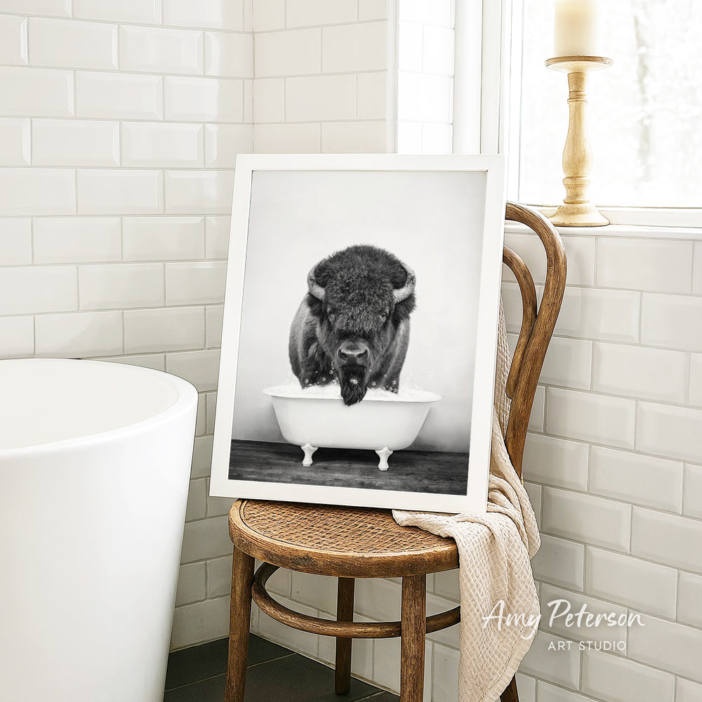 A framed black and white photograph of a bison sitting in a bathtub is displayed on a wooden chair in a bathroom setting.
