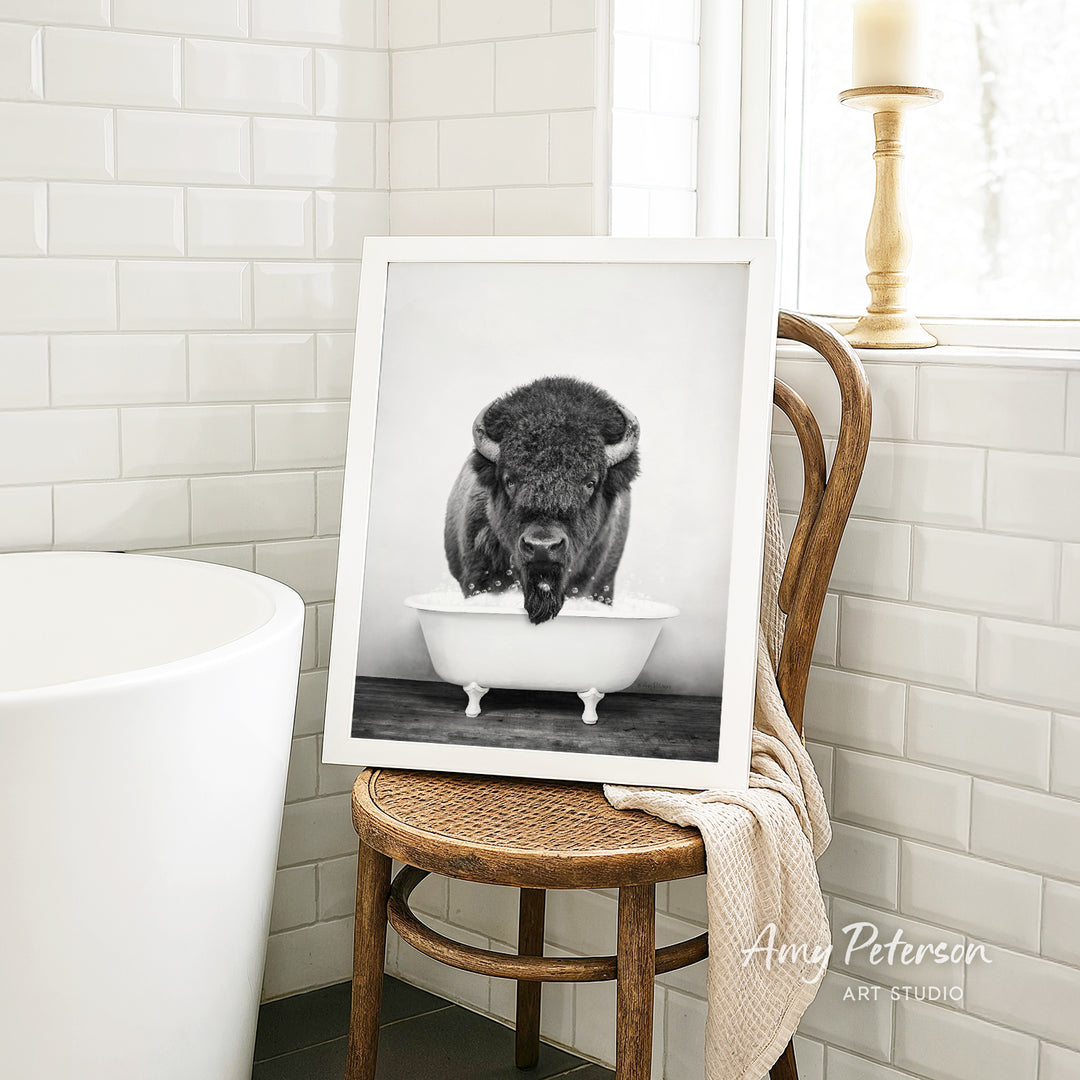 A framed black and white photograph of a bison sitting in a bathtub is displayed on a wooden chair in a bathroom setting.