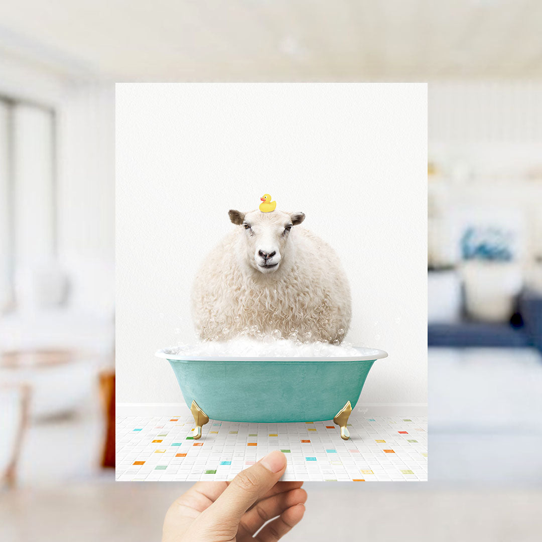 A hand holds up a framed image of a fluffy white sheep sitting in a blue bathtub filled with bubbles, with a yellow rubber duck perched on its head.