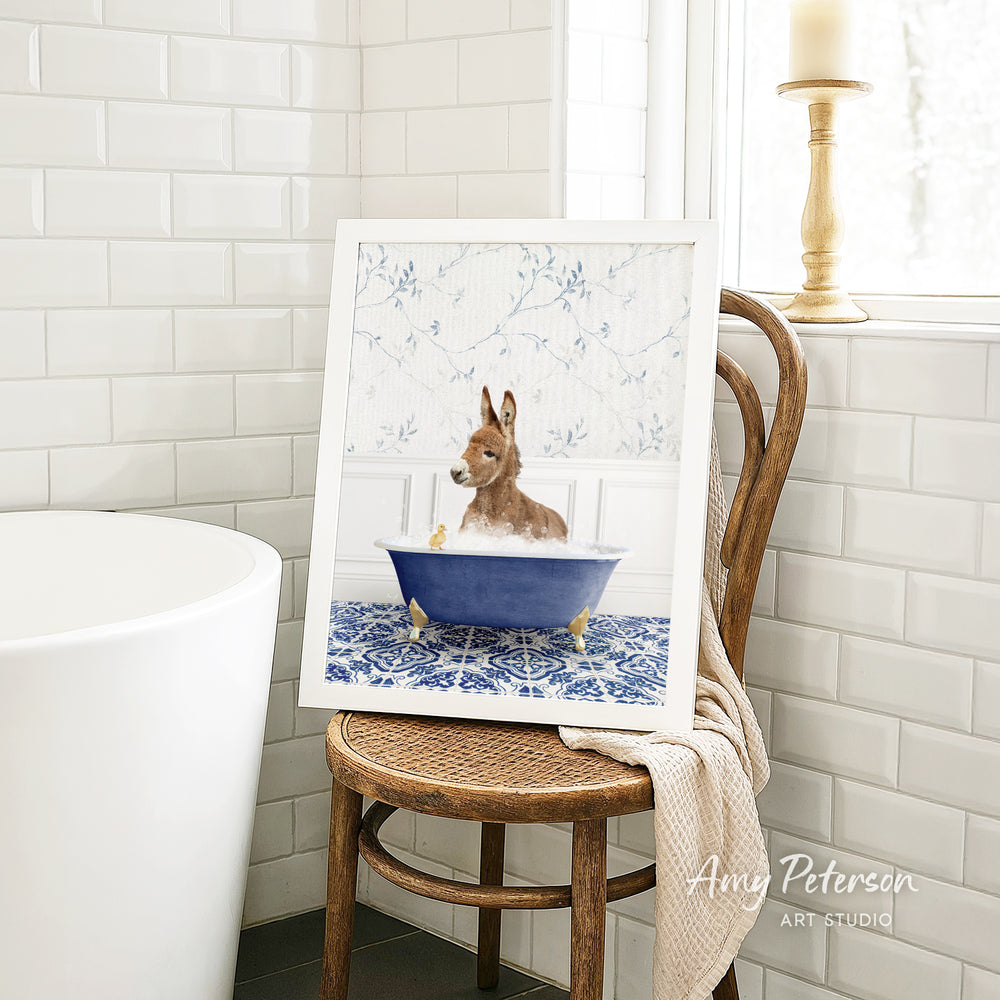 A framed image of a brown rabbit sitting in a blue bathtub, surrounded by a white floral pattern, is displayed on a wooden chair in a bathroom setting.