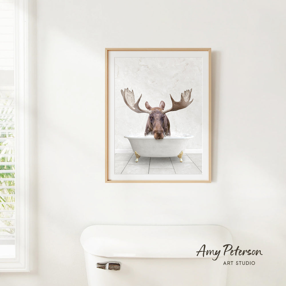 A framed image of a moose sitting in a bathtub, with its large antlers raised, is hanging on a white wall.