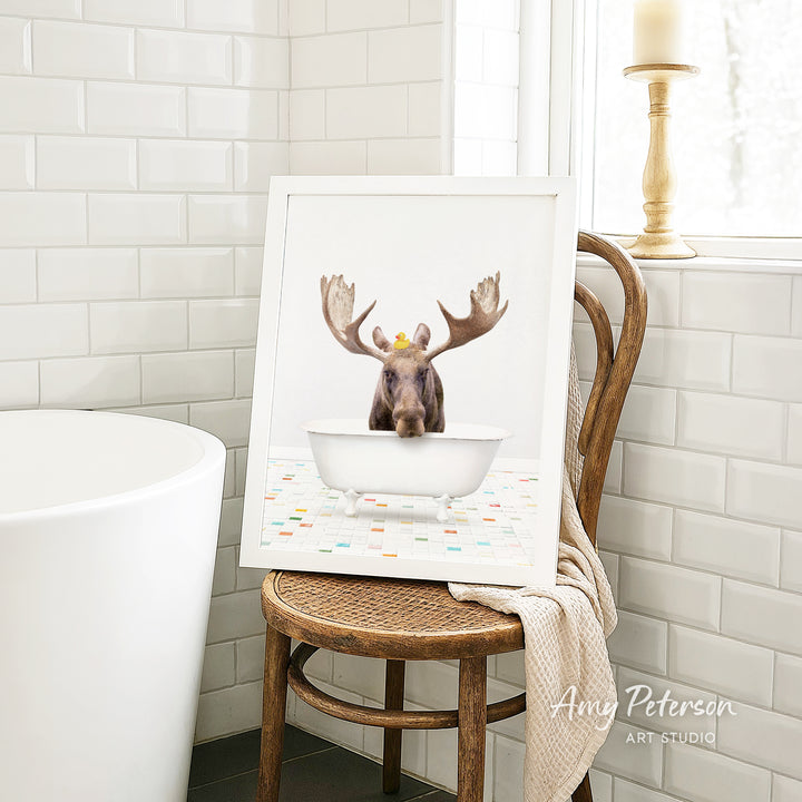 A framed image of a moose with antlers sitting in a bathtub, surrounded by colorful tiles, is displayed on a wooden chair in a bathroom.