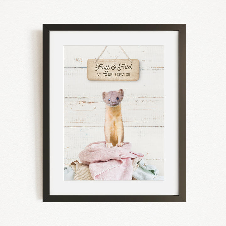 A framed image of a small animal, likely a cat, sitting on a pile of clothes and towels, with a wooden sign above that says "Fluffy & Fold at your service".