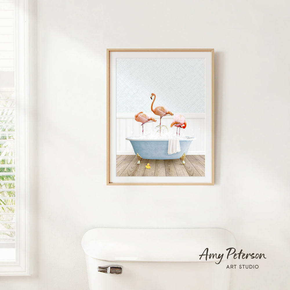 A framed print of three flamingos sitting in a bathtub is hanging on a white wall in a bathroom.