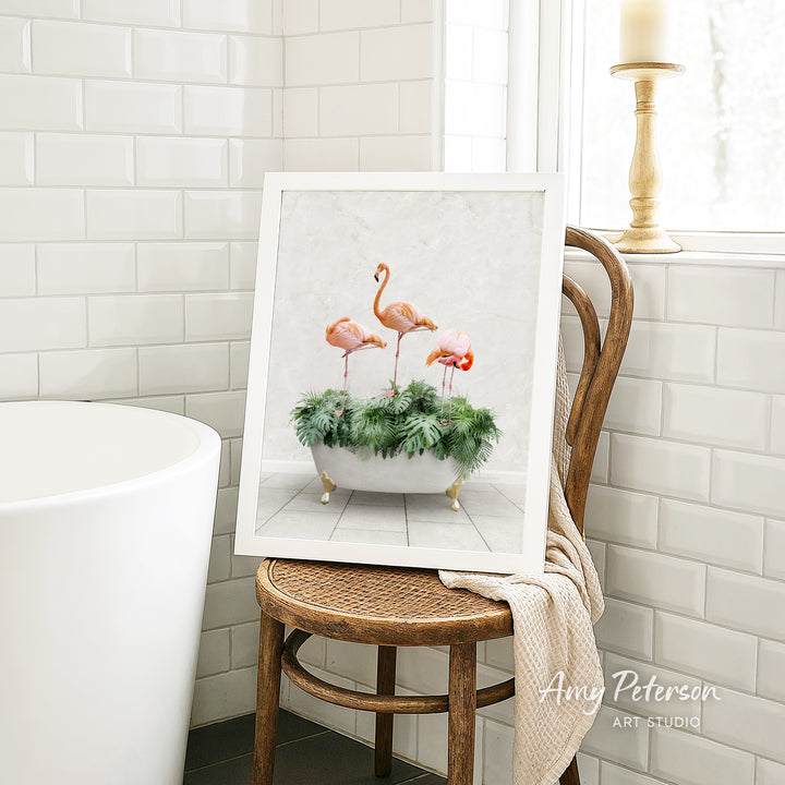 A framed print of three flamingos standing in a bathtub filled with green plants is displayed on a wooden chair in a bathroom setting.