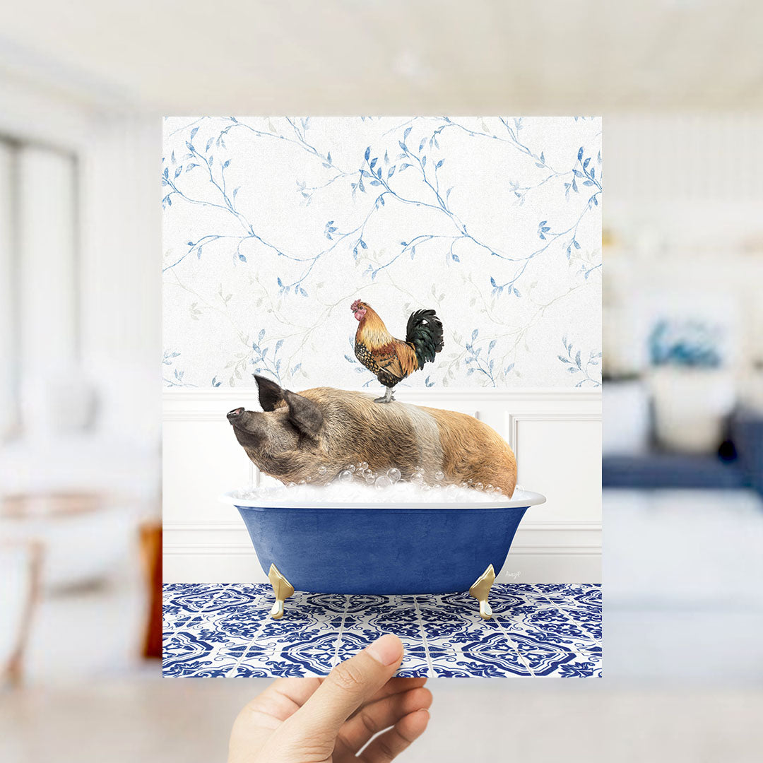 A hand holds up a framed image depicting a pig sitting in a blue bathtub, with a rooster perched on its back.