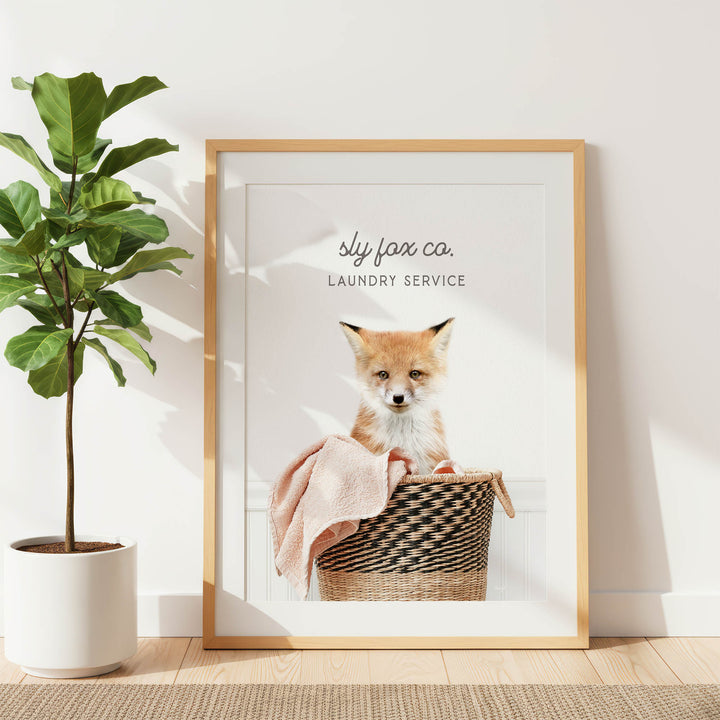 A framed image of a fox sitting in a laundry basket, with a towel draped over it, is displayed on a wall next to a potted plant.