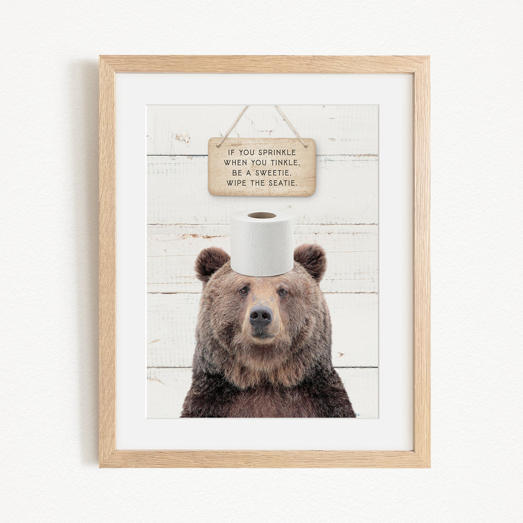 A framed image of a brown bear wearing a toilet paper roll on its head, with a wooden sign above it that reads "If you sprinkle when you twinkle, be a sweetie, wipe the seattle."