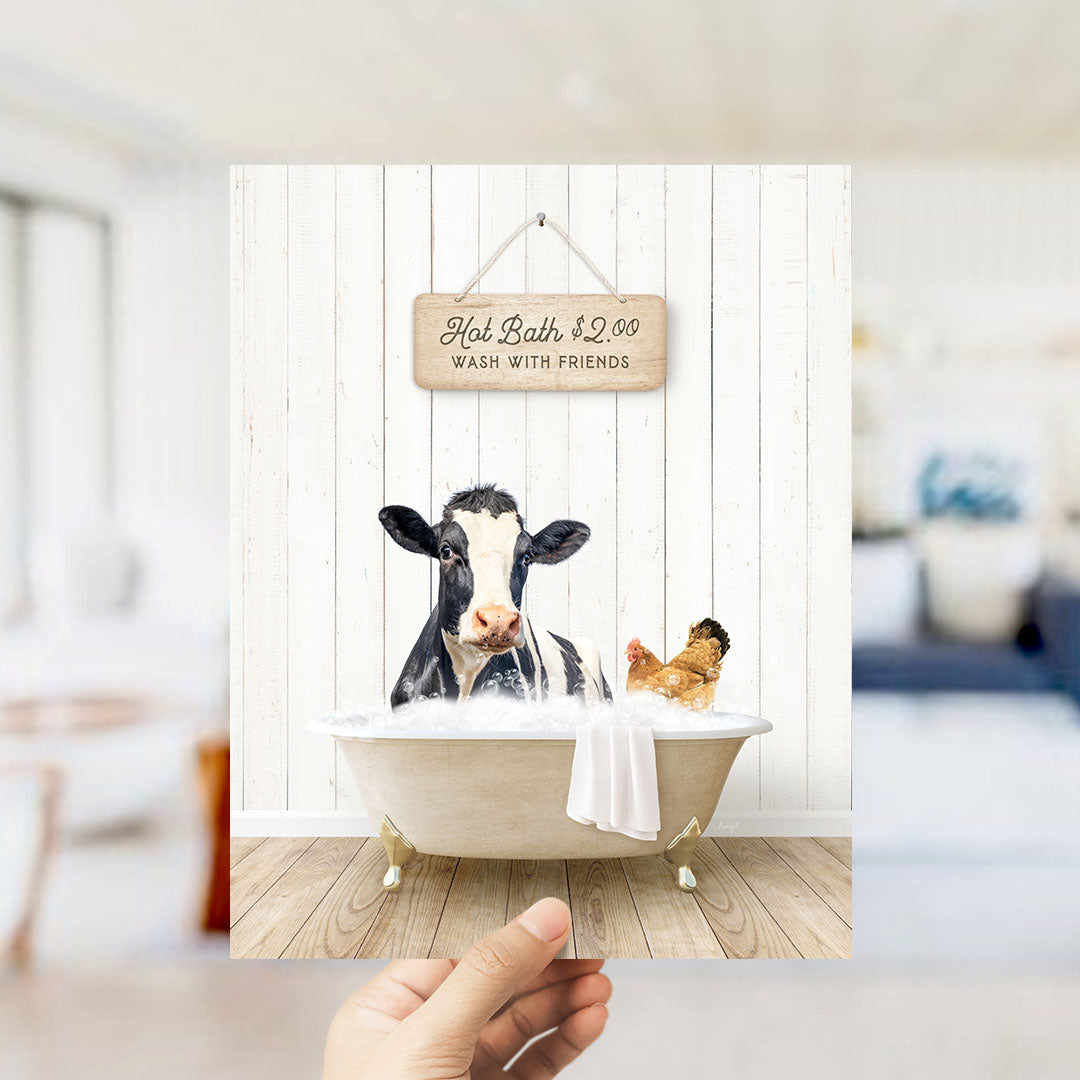 A hand holds up a framed image depicting a cow and a chicken relaxing in a bathtub, with a sign above them advertising a hot bath for $2.00.