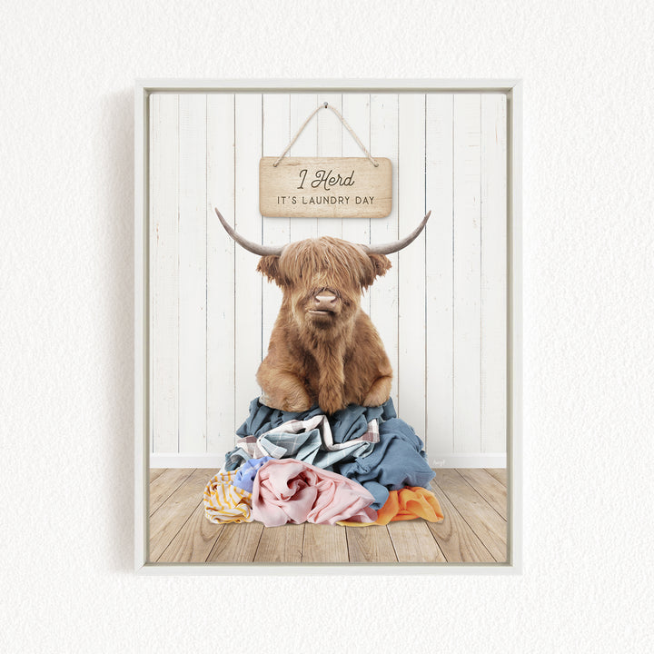 A brown cow with horns is sitting on a pile of clothes, with a sign above it that says "I herd it's laundry day".