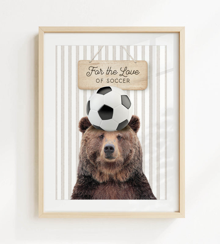 A framed image of a brown bear with a soccer ball on its head, accompanied by a wooden sign that reads "For the Love of Soccer".