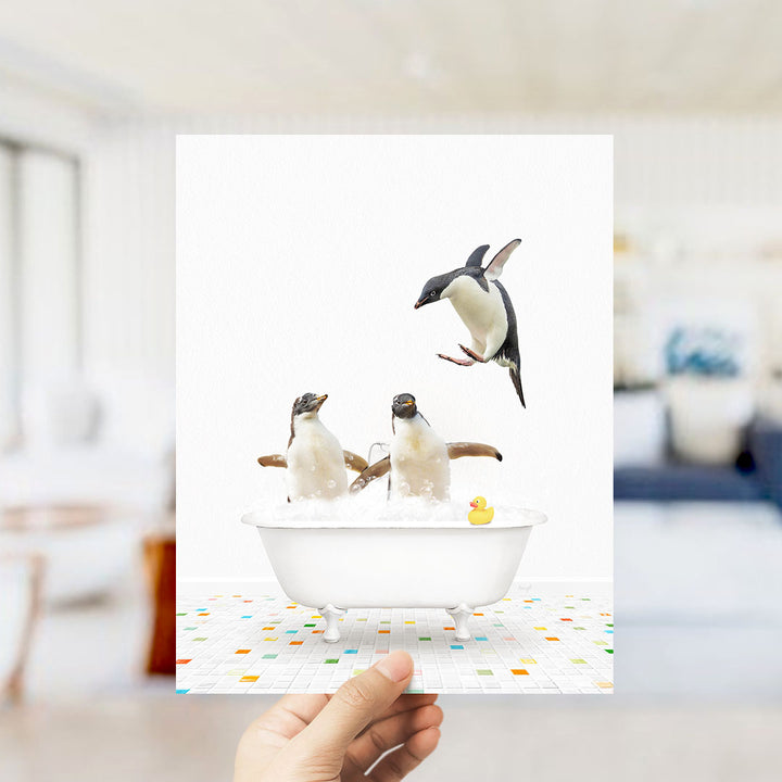 A hand holds up a framed image depicting two penguins sitting in a bathtub, with one penguin flying towards the other.