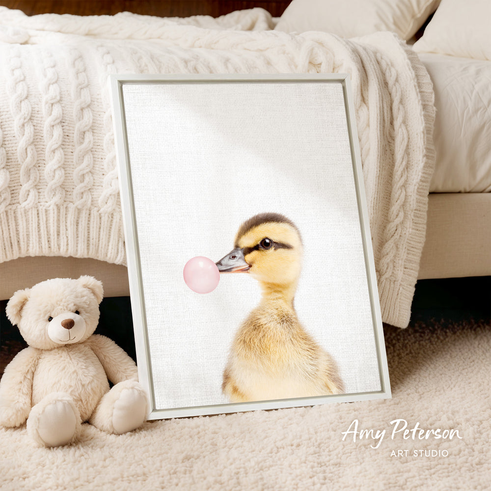 A framed image of a duck blowing a bubble gum bubble is displayed on a white bedspread, accompanied by a teddy bear on the floor.