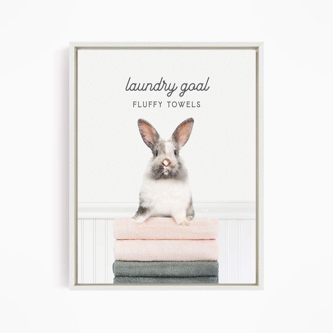 A fluffy white rabbit sitting on a stack of towels, with the text "laundry goal FLUFFY TOWELS" displayed above the rabbit.