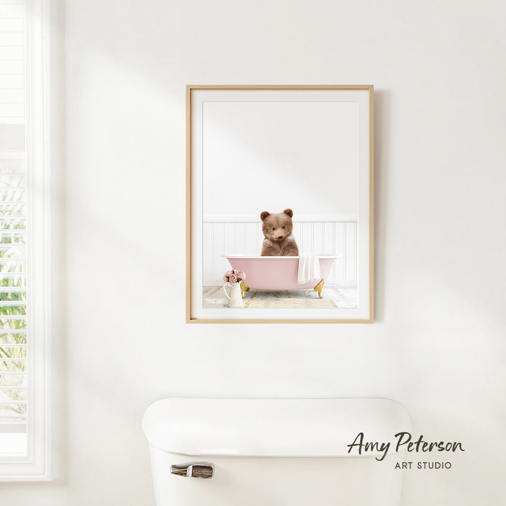 A framed image of a brown bear sitting in a pink bathtub, with a vase of flowers and a white toilet in the foreground.