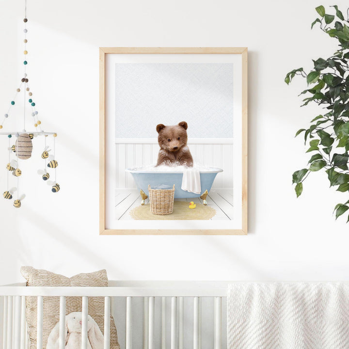 A framed image of a brown bear sitting in a blue bathtub, surrounded by a basket of bath toys and a white crib in a nursery setting.
