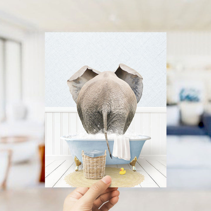 A hand holds up a framed image depicting an elephant standing in a bathtub filled with water, surrounded by a yellow rubber duck and a white towel.