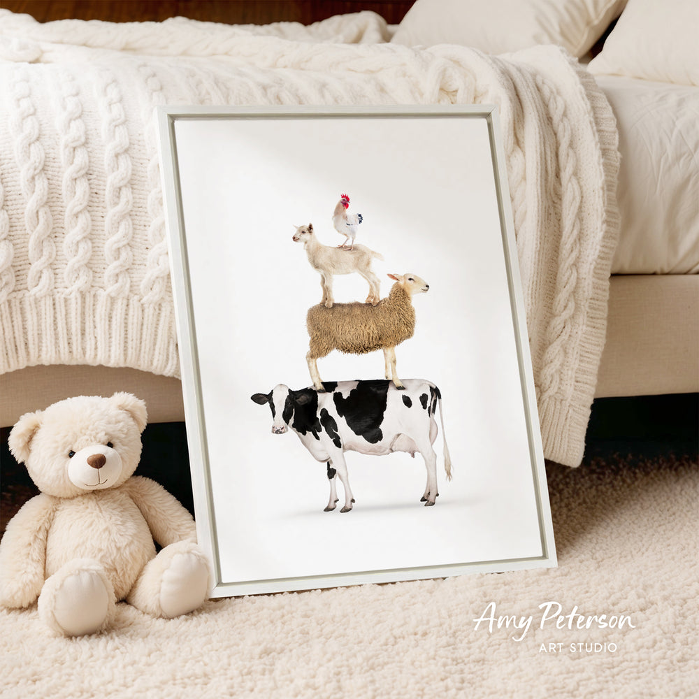 A framed illustration of a cow, sheep, and chicken stacked on top of each other, with a teddy bear sitting next to it on a white blanket.