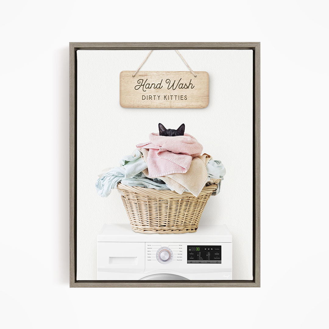 A wooden sign with the text "Hand Wash Dirty Kittens" hangs above a wicker basket filled with clothes, with a black cat peeking out from the basket.