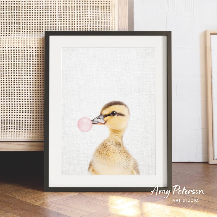 A framed photograph of a duck blowing a bubble gum bubble.
