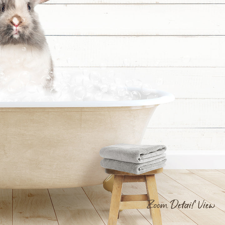 A fluffy gray rabbit sitting in a bathtub filled with bubbles, with a wooden stool and towels nearby.