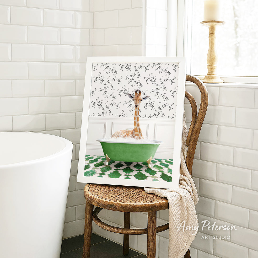 A framed image of a giraffe sitting in a green bathtub, surrounded by a floral pattern, is displayed on a wooden chair in a bathroom setting.