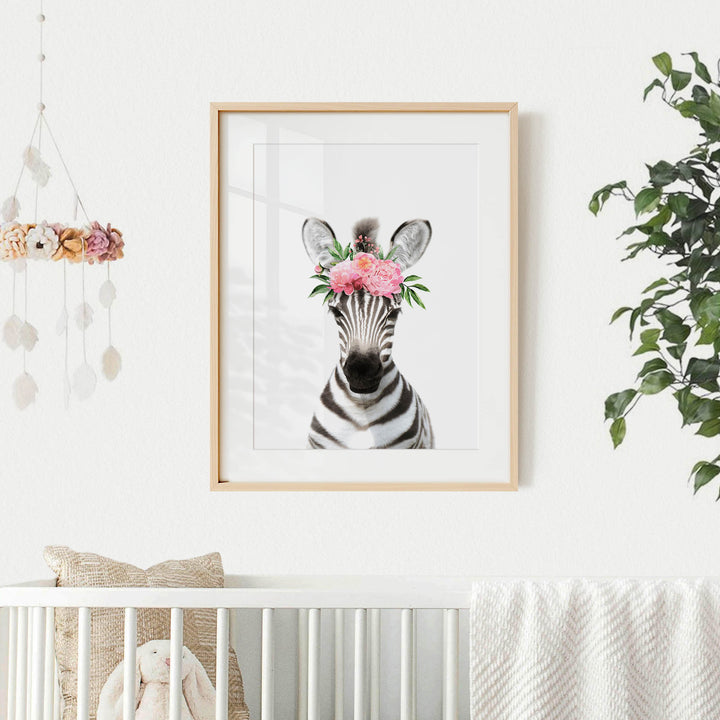 A framed print of a zebra wearing a flower crown is displayed on a white wall, with a crib and a hanging mobile with flowers in the background.
