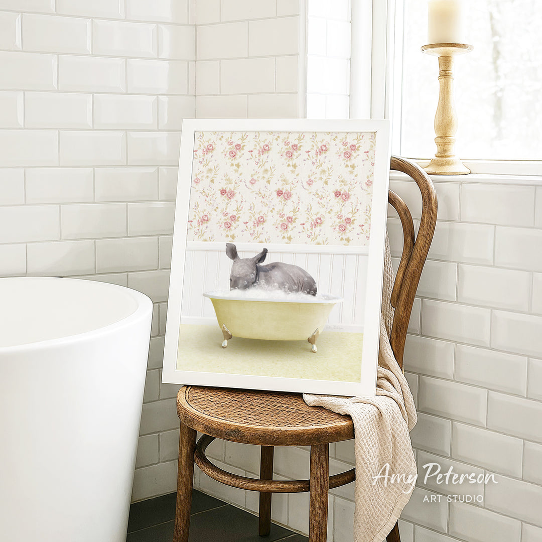 A framed image of a gray rabbit sitting in a yellow bathtub, surrounded by a floral pattern, is displayed on a wooden chair in a bathroom setting.