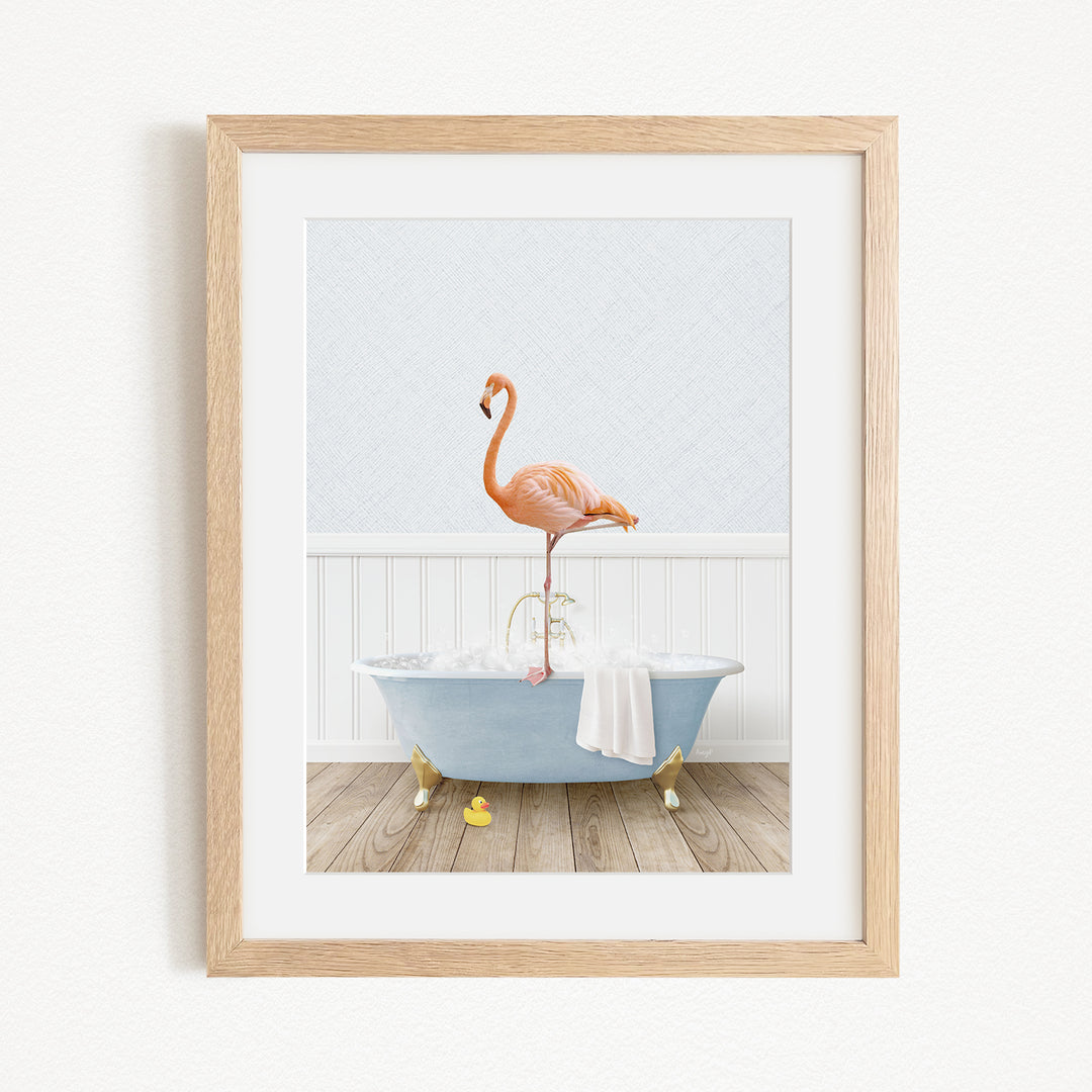 A framed image depicting a pink flamingo standing on a blue bathtub, with a white towel and yellow rubber duckies on the floor.