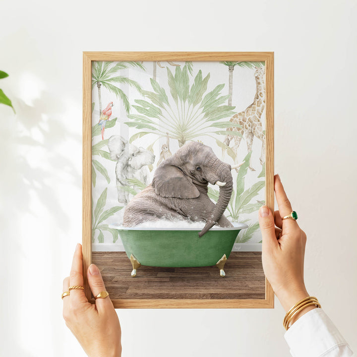 A person's hands are holding a framed picture of an elephant sitting in a green bathtub, surrounded by tropical foliage.