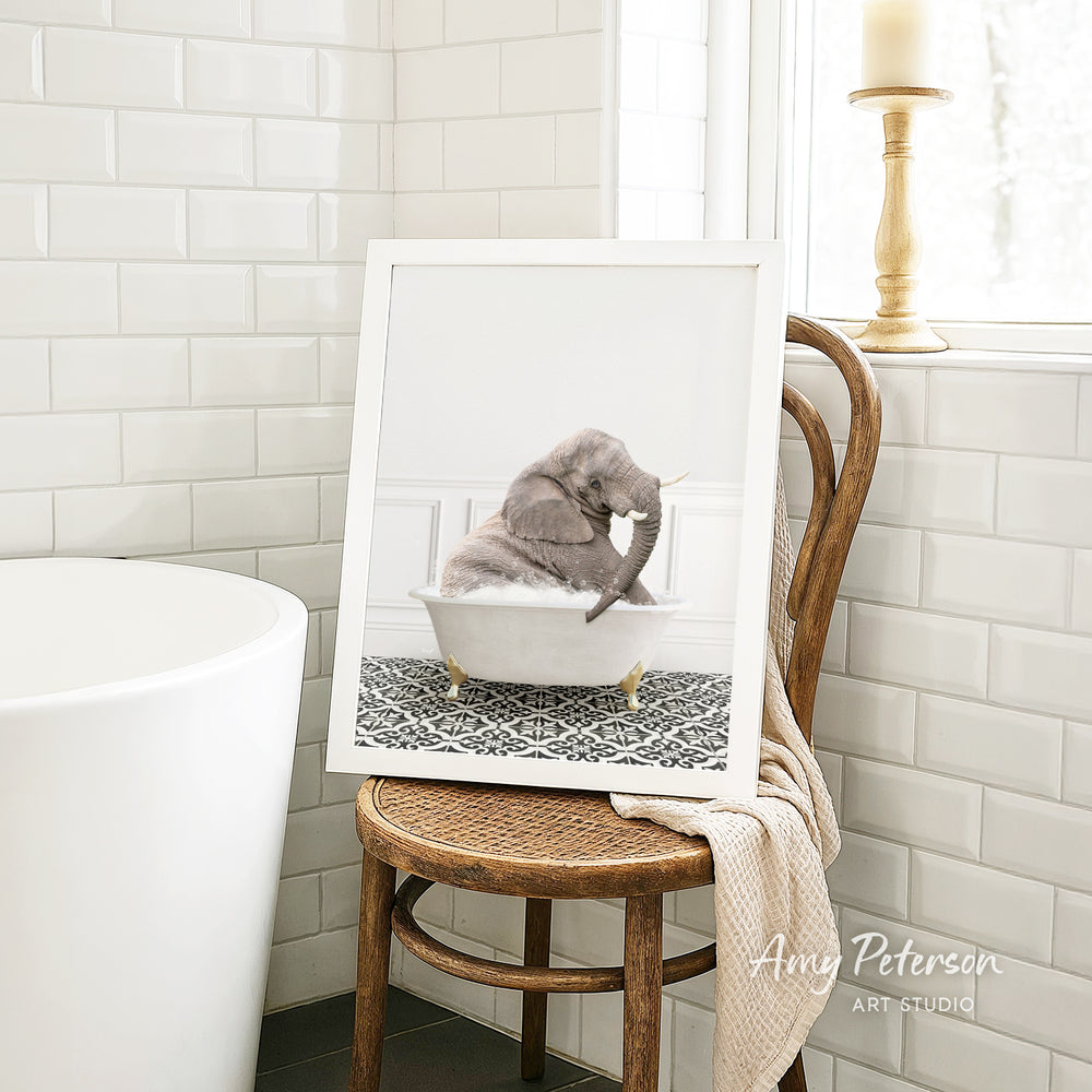 A framed photograph of a gray elephant sitting in a white bathtub, with a towel draped over the back of a wooden chair in a bathroom setting.