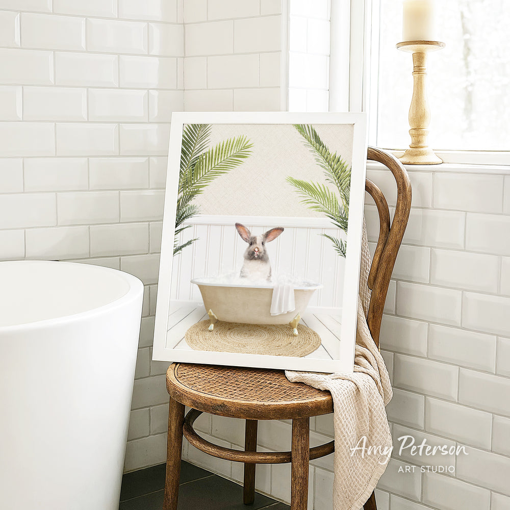 A framed image of a pig sitting in a bathtub, surrounded by palm leaves, is displayed on a wooden chair in a bathroom setting.