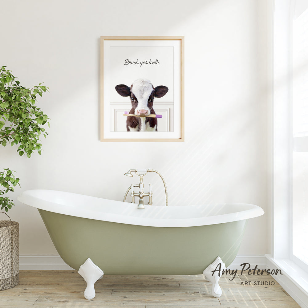A framed image of a cow with a toothbrush in its mouth is displayed on a white wall above a green clawfoot bathtub in a bright, airy bathroom.