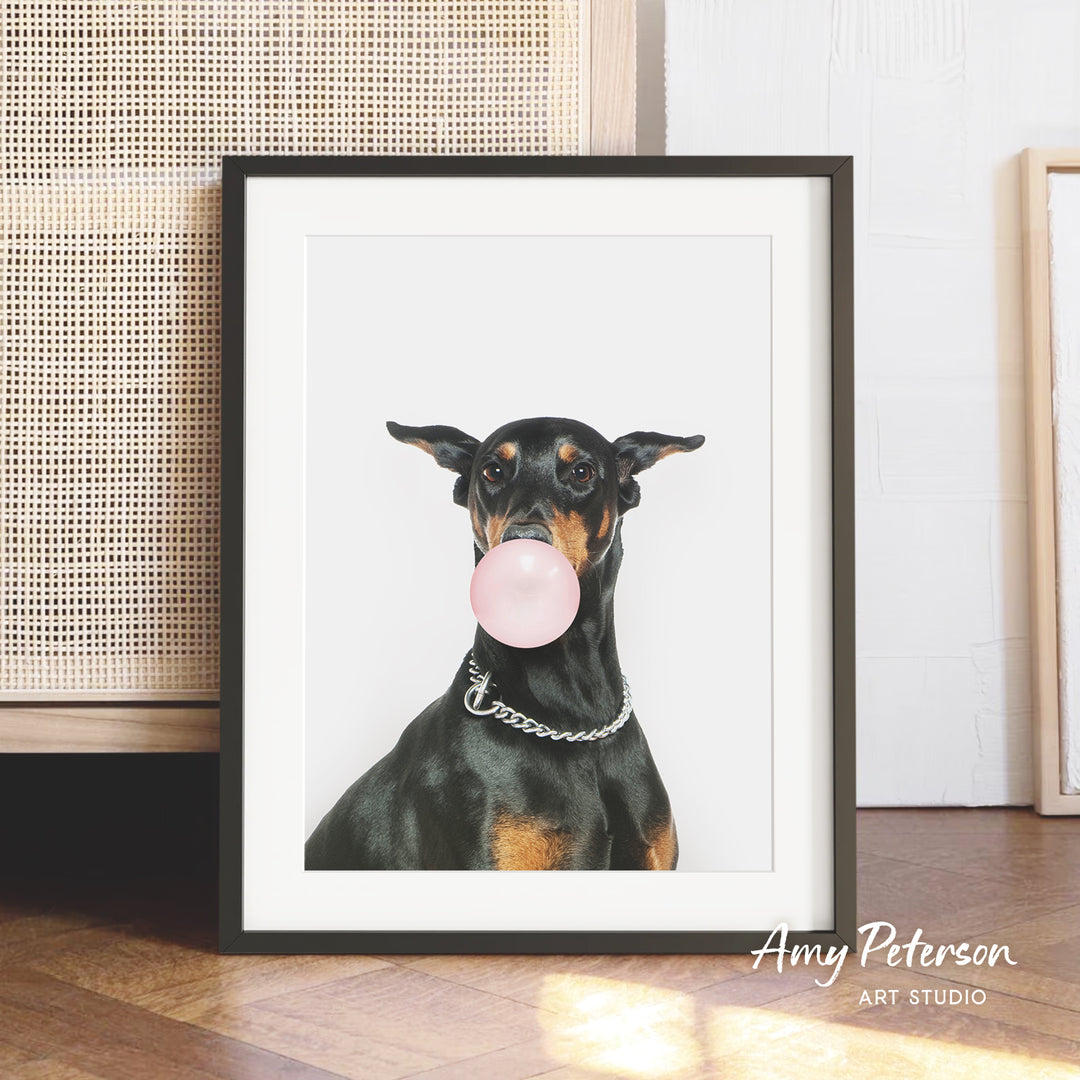 A black and tan dog is sitting on a wooden floor, holding a pink bubble gum bubble in its mouth.