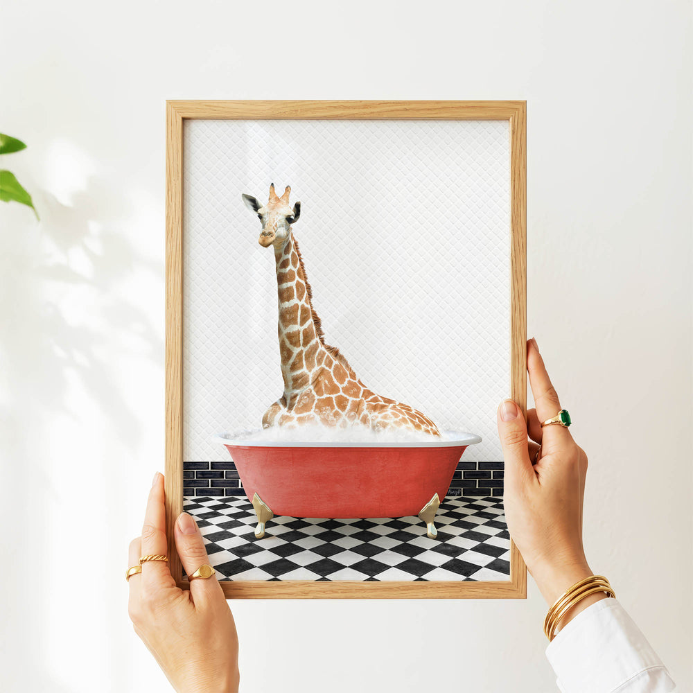 A person's hands are holding a framed picture of a giraffe sitting in a red bathtub on a black and white checkered floor.