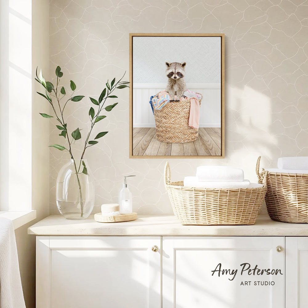 a raccoon sitting in a woven basket, which is placed on a white countertop. The raccoon is surrounded by various items, including a vase with green leaves, a soap dispenser, and a towel. The backdrop is a beige wall with a framed picture of a raccoon.
