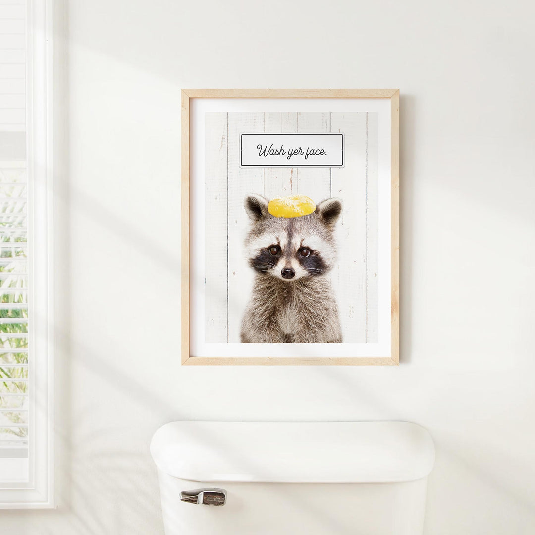 A framed picture of a raccoon wearing a yellow hat with the text "Wash your face" written on it, hanging on a white wall above a toilet.