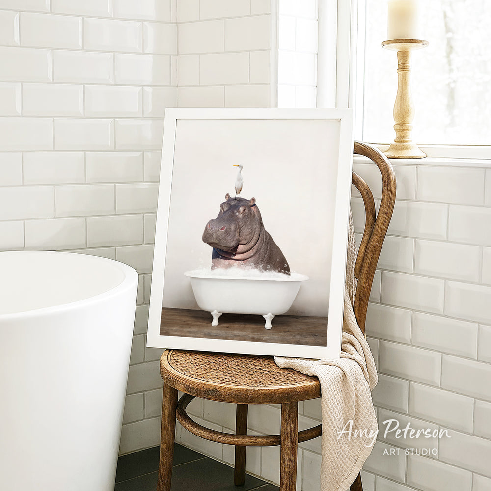 A framed photograph of a hippopotamus sitting in a bathtub, with a bird perched on its head, is displayed on a wooden chair in a bathroom setting.
