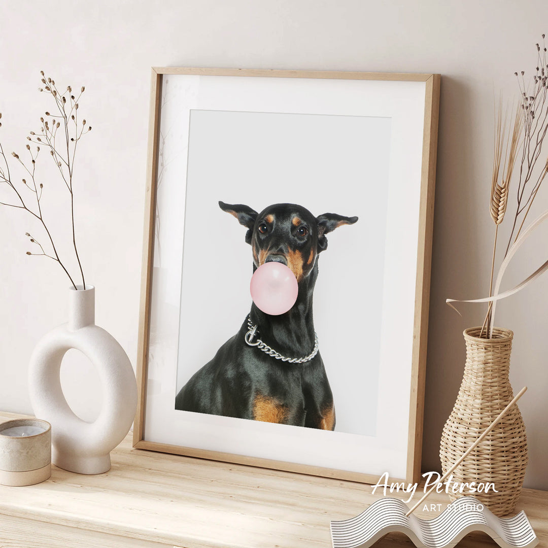 A framed photograph of a Doberman Pinscher dog blowing a bubble with a pink bubble gum bubble, accompanied by decorative vases and a text that reads " Amy Peterson ART STUDIO".