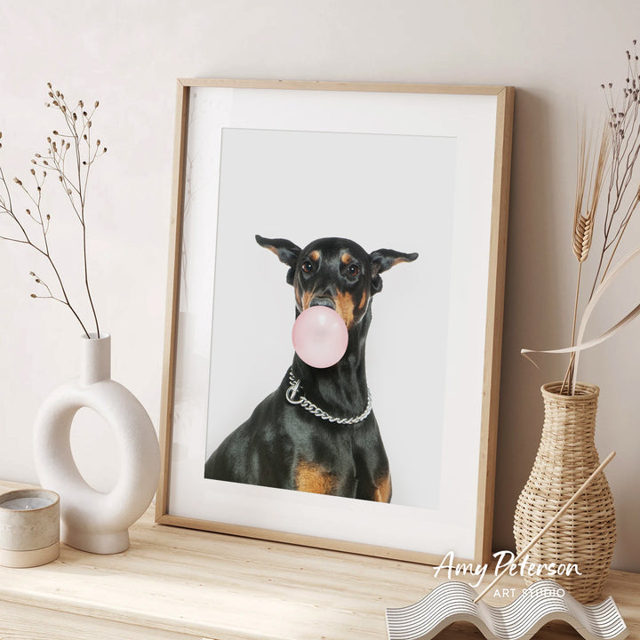 A framed photograph of a Doberman Pinscher dog blowing a bubble with a pink bubble gum bubble, accompanied by decorative vases and a text that reads " Amy Peterson ART STUDIO".