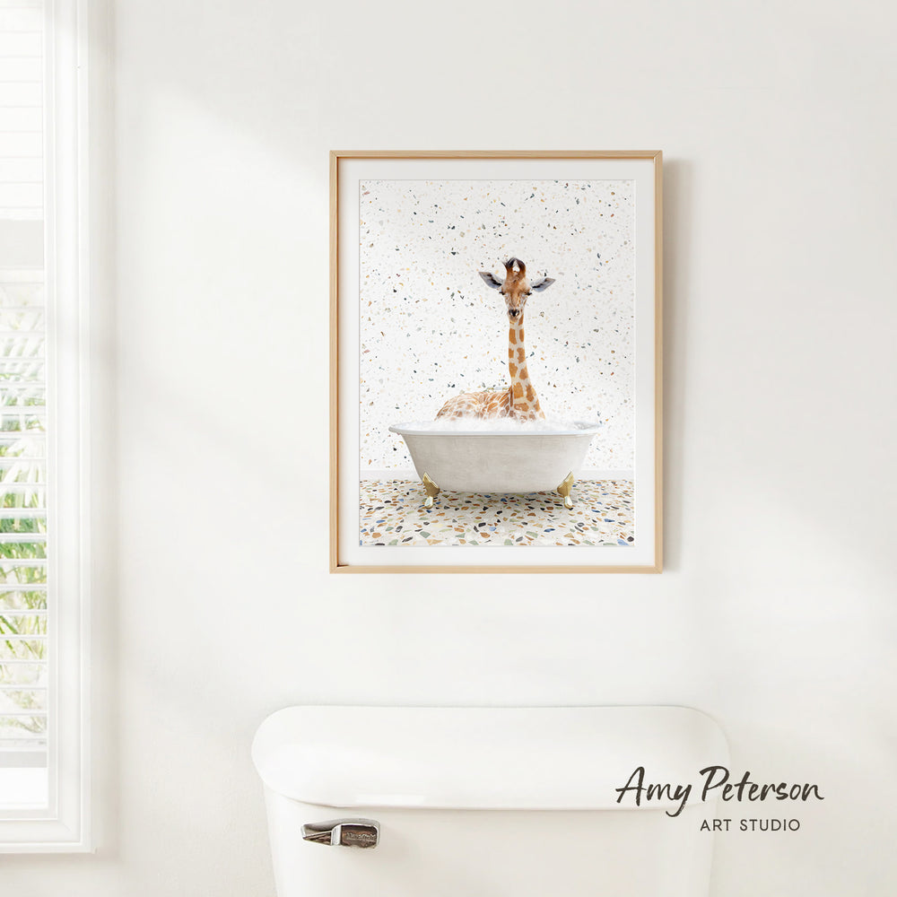 A framed painting of a giraffe sitting in a bathtub, surrounded by confetti, hangs on a white wall.