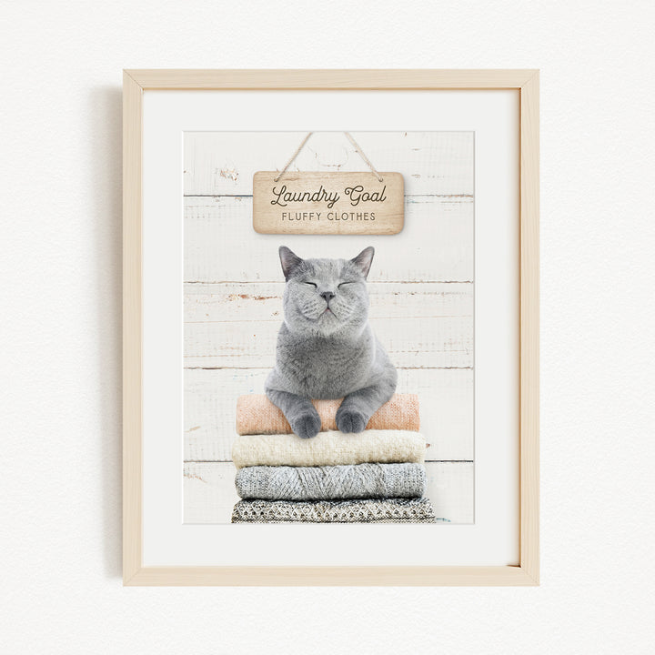 A framed image of a gray cat sitting on a stack of folded clothes, with a wooden sign above that says "Laundry goal fluffy clothes".