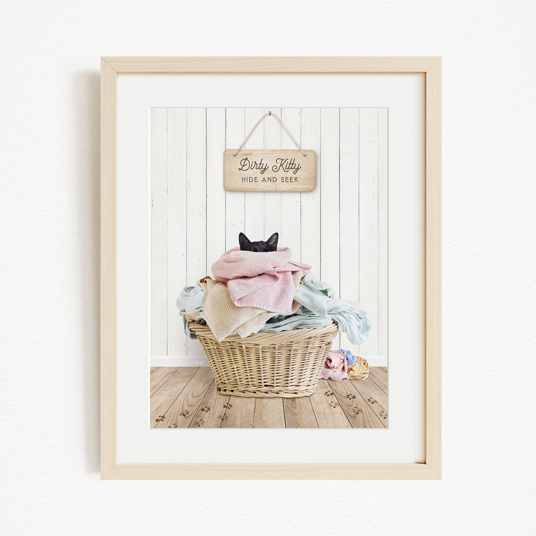 A framed photograph of a cat sitting in a basket filled with laundry, with a sign above the basket that reads "Dirty Kitty HIDE AND SEEK".